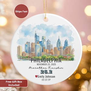 May include: A round, white ceramic ornament featuring a watercolor illustration of the Philadelphia skyline. The ornament includes the text "PHILADELPHIA," "NOVEMBER 23, 2025," "Marathon Finisher," "26.2," a name, and a time. A gold string is attached for hanging.