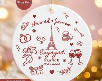 May include: White ceramic ornament with a gold string, featuring a red design of the Eiffel Tower, hearts, and the words "Hannah & James" and "Engaged France 10.24.2025". Includes illustrations of rings, macarons, and a cupid.