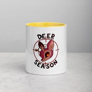 Puede incluir: Taza de cerámica blanca con interior amarillo. La taza presenta una cabeza de ciervo de dibujos animados dentro de una diana, con las palabras "DEER SEASON" en letras negras. El ciervo es marrón y rojo.