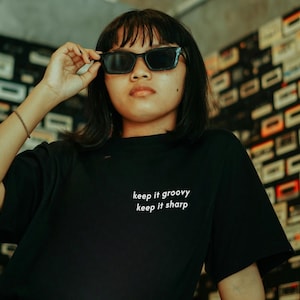 Puede incluir: Una persona con gafas de sol negras y una camiseta negra. La camiseta tiene las palabras "keep it groovy keep it sharp" impresas en blanco. El fondo está desenfocado.