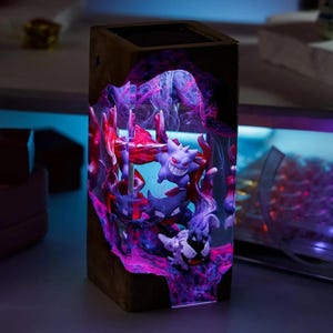 Peut inclure: Lampe rectangulaire en bois avec une sc&egrave;ne illumin&eacute;e mettant en vedette des personnages Pok&eacute;mon. L'int&eacute;rieur brille de teintes violettes et roses, pr&eacute;sentant les figurines dans une mati&egrave;re semblable &agrave; de la r&eacute;sine. Un objet d&eacute;coratif unique.