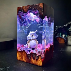 Peut inclure: Une &oelig;uvre d'art en r&eacute;sine rectangulaire repr&eacute;sentant un personnage de Cubone. L'&oelig;uvre a une base en bois avec un design color&eacute; et superpos&eacute;. L'int&eacute;rieur pr&eacute;sente un effet n&eacute;buleux violet et bleu, avec le personnage de Cubone au centre.