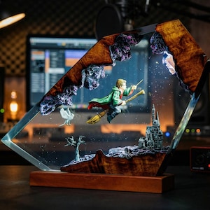 May include: A decorative resin sculpture featuring a miniature figure on a broomstick, a castle, and a bird. The sculpture is encased in clear resin with a wooden base and accents. The background includes a computer screen and studio equipment.