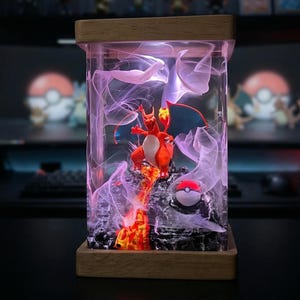 May include: A rectangular resin lamp featuring a Charizard figure, flames, and a Poké Ball. The lamp has a wooden base and top, with a clear resin body filled with purple and white wisps. The Charizard is orange, red, and white.