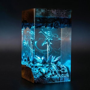 May include: A rectangular resin art piece with a glowing blue interior. The piece features a dark wooden base and top, with a detailed, textured blue landscape and crystalline structures inside. The overall effect is a luminous, otherworldly display.