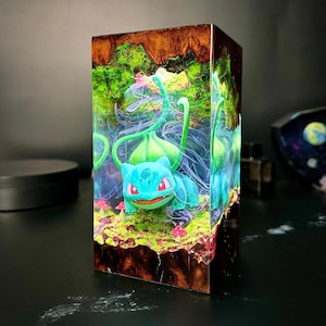 Peut inclure: Bloc de r&eacute;sine repr&eacute;sentant un personnage de Bulbasaur dans une sc&egrave;ne de for&ecirc;t miniature. La figurine est bleue et verte avec des yeux rouges, entour&eacute;e de feuillage vert, de champignons et d'une base rocheuse brune. Le bloc a un ext&eacute;rieur brun fonc&eacute;.