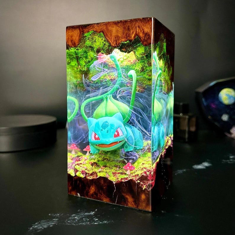 Peut inclure: Bloc de r&eacute;sine repr&eacute;sentant un personnage de Bulbasaur dans une sc&egrave;ne de for&ecirc;t miniature. La figurine est bleue et verte avec des yeux rouges, entour&eacute;e de feuillage vert, de champignons et d'une base rocheuse brune. Le bloc a un ext&eacute;rieur brun fonc&eacute;.