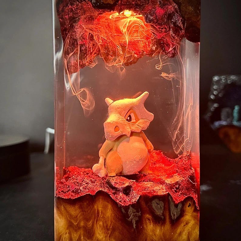 Peut inclure: Un bloc de r&eacute;sine transparent renferme une figurine Cubone orange clair, sur un fond rouge et orange flamboyant. La r&eacute;sine est &eacute;clair&eacute;e par une lumi&egrave;re interne, cr&eacute;ant un effet lumineux. La figurine Cubone est le point central de cette &oelig;uvre d'art unique.