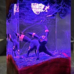 Peut inclure: Une sculpture en r&eacute;sine repr&eacute;sentant un Umbreon noir avec des yeux et des accents rouges, sur fond de tourbillons violets et blancs. La sculpture est enferm&eacute;e dans un bloc de r&eacute;sine transparent, avec une base rouge et marron.