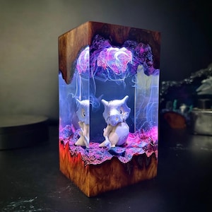 Peut inclure: Bloc de bois artisanal avec un int&eacute;rieur en r&eacute;sine, mettant en vedette deux figurines de Cubone. La r&eacute;sine est illumin&eacute;e de teintes bleues et violettes, cr&eacute;ant un effet lumineux. L'ext&eacute;rieur en bois a un aspect naturel et textur&eacute;, de couleur marron chaud.