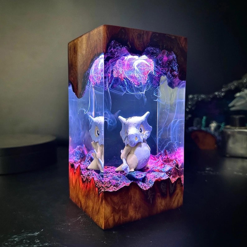 Peut inclure: Bloc de bois artisanal avec un int&eacute;rieur en r&eacute;sine, mettant en vedette deux figurines de Cubone. La r&eacute;sine est illumin&eacute;e de teintes bleues et violettes, cr&eacute;ant un effet lumineux. L'ext&eacute;rieur en bois a un aspect naturel et textur&eacute;, de couleur marron chaud.