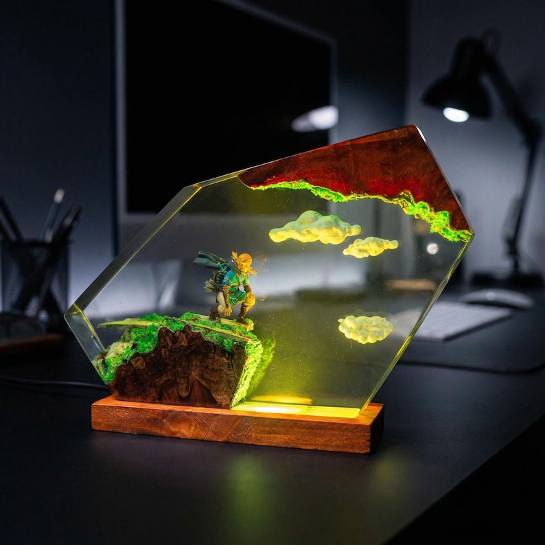 Puede incluir: L&aacute;mpara decorativa con una figura en miniatura de pie sobre un paisaje verde y marr&oacute;n, encerrada en un bloque de resina transparente y angular. La resina incluye nubes amarillas y una parte superior roja y verde. La l&aacute;mpara se asienta sobre una base de madera.
