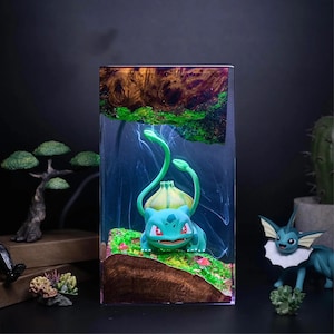 Peut inclure: Bloc de r&eacute;sine transparent avec une figurine de Bulbasaur, pos&eacute;e sur un paysage miniature avec de la mousse verte et de la terre brune. La r&eacute;sine a une section sup&eacute;rieure en forme de bois avec des accents verts. D'autres figurines d'un bonsa&iuml; et d'un Vaporeon sont visibles.