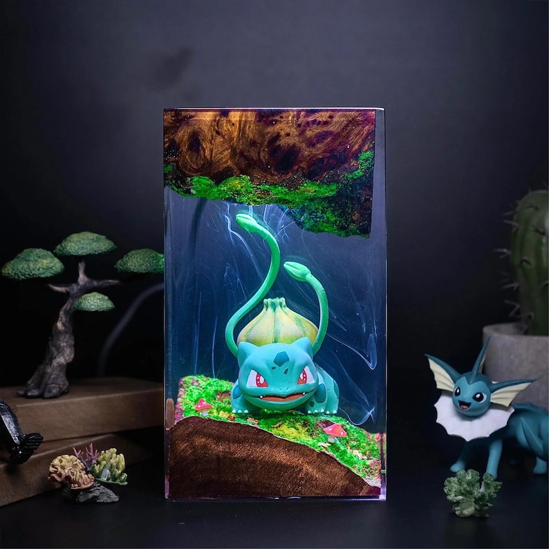 Peut inclure: Bloc de r&eacute;sine transparent avec une figurine de Bulbasaur, pos&eacute;e sur un paysage miniature avec de la mousse verte et de la terre brune. La r&eacute;sine a une section sup&eacute;rieure en forme de bois avec des accents verts. D'autres figurines d'un bonsa&iuml; et d'un Vaporeon sont visibles.
