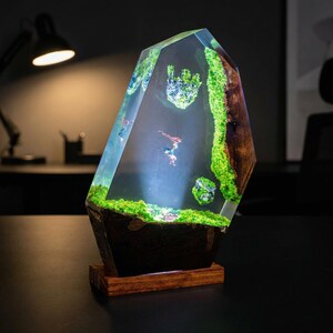 Puede incluir: L&aacute;mpara decorativa de resina con un dise&ntilde;o &uacute;nico. La l&aacute;mpara presenta una forma clara y facetada con un paisaje interior de musgo verde, figuras en miniatura y una base de madera. La l&aacute;mpara est&aacute; iluminada, creando una exhibici&oacute;n cautivadora.