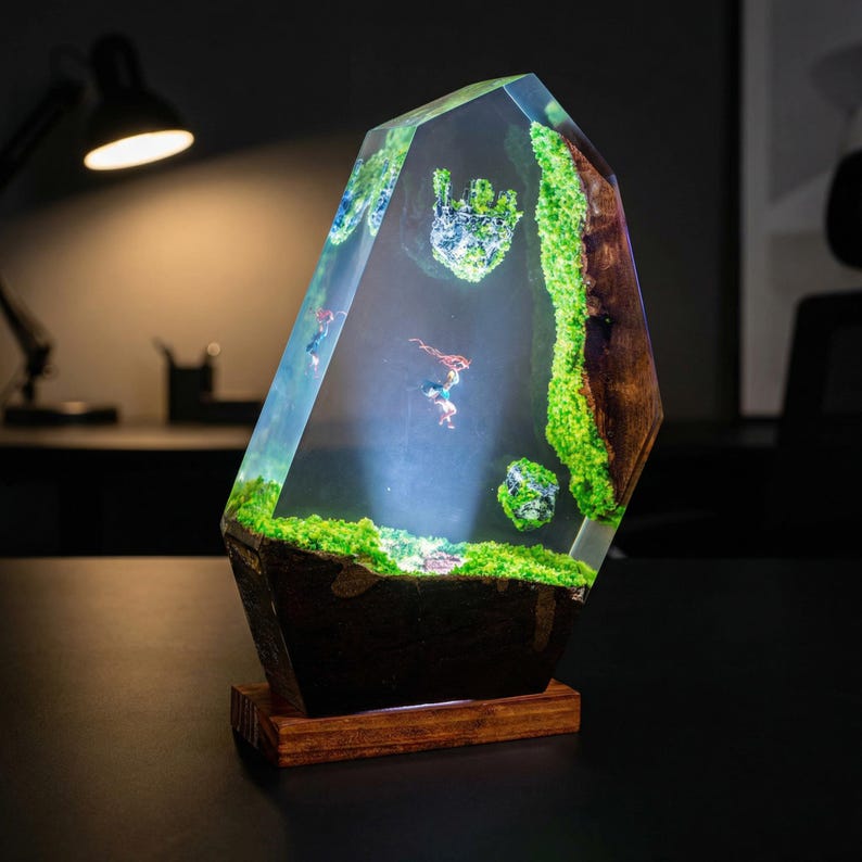 Puede incluir: L&aacute;mpara decorativa de resina con un dise&ntilde;o &uacute;nico. La l&aacute;mpara presenta una forma clara y facetada con un paisaje interior de musgo verde, figuras en miniatura y una base de madera. La l&aacute;mpara est&aacute; iluminada, creando una exhibici&oacute;n cautivadora.
