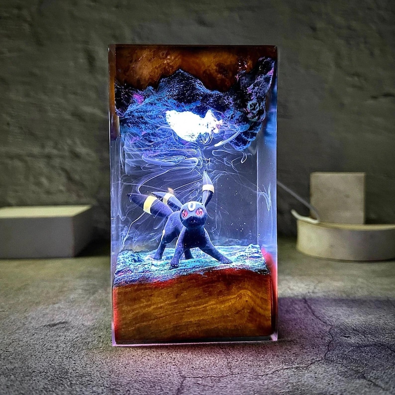 Peut inclure: Une pi&egrave;ce d'art en r&eacute;sine rectangulaire repr&eacute;sentant un Umbreon noir avec des accents jaunes, sur fond d'&eacute;l&eacute;ments abstraits bleus et bruns. La base a l'apparence du bois et la pi&egrave;ce est &eacute;clair&eacute;e d'une douce lueur.