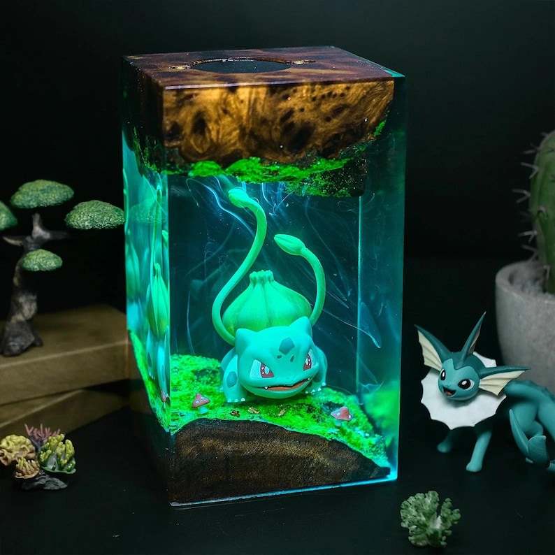 Peut inclure: Une sculpture en r&eacute;sine rectangulaire repr&eacute;sentant un Bulbasaur dans un paysage miniature. La sculpture a un dessus en bois et un int&eacute;rieur bleu brillant. Le Bulbasaur est vert, avec des yeux rouges, et est pos&eacute; sur de la mousse verte.