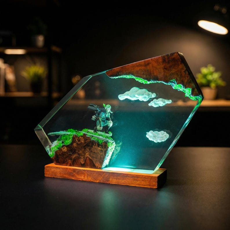 Puede incluir: Escultura decorativa de resina con una figura en miniatura sobre un acantilado, con nubes y una base de madera. La escultura tiene un dise&ntilde;o claro y angular con un paisaje verde y marr&oacute;n, y un fondo azul claro.