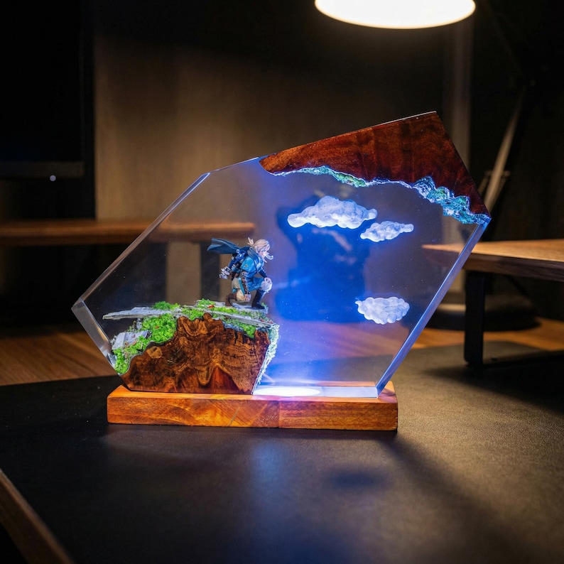 Puede incluir: Una escultura decorativa de resina con una figura en miniatura sobre un paisaje rocoso con musgo verde y nubes blancas. La escultura est&aacute; encapsulada en resina transparente con una base de madera y una luz azul.
