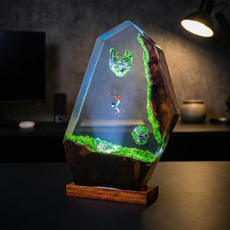 Puede incluir: Escultura de resina transparente con base de madera. En el interior, un paisaje en miniatura con musgo verde, un elemento de madera marr&oacute;n y una peque&ntilde;a figura. La escultura tiene forma geom&eacute;trica y est&aacute; iluminada.