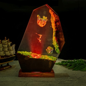 Puede incluir: L&aacute;mpara decorativa de resina con un dise&ntilde;o &uacute;nico. La l&aacute;mpara presenta una forma geom&eacute;trica con paisajes en miniatura incrustados en tonos verdes, naranjas y rojos. Se apoya sobre una base de madera, creando un brillo ambiental c&aacute;lido.