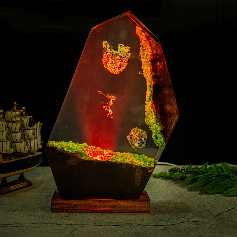 Puede incluir: L&aacute;mpara decorativa de resina con un dise&ntilde;o &uacute;nico. La l&aacute;mpara presenta una forma geom&eacute;trica con paisajes en miniatura incrustados en tonos verdes, naranjas y rojos. Se apoya sobre una base de madera, creando un brillo ambiental c&aacute;lido.