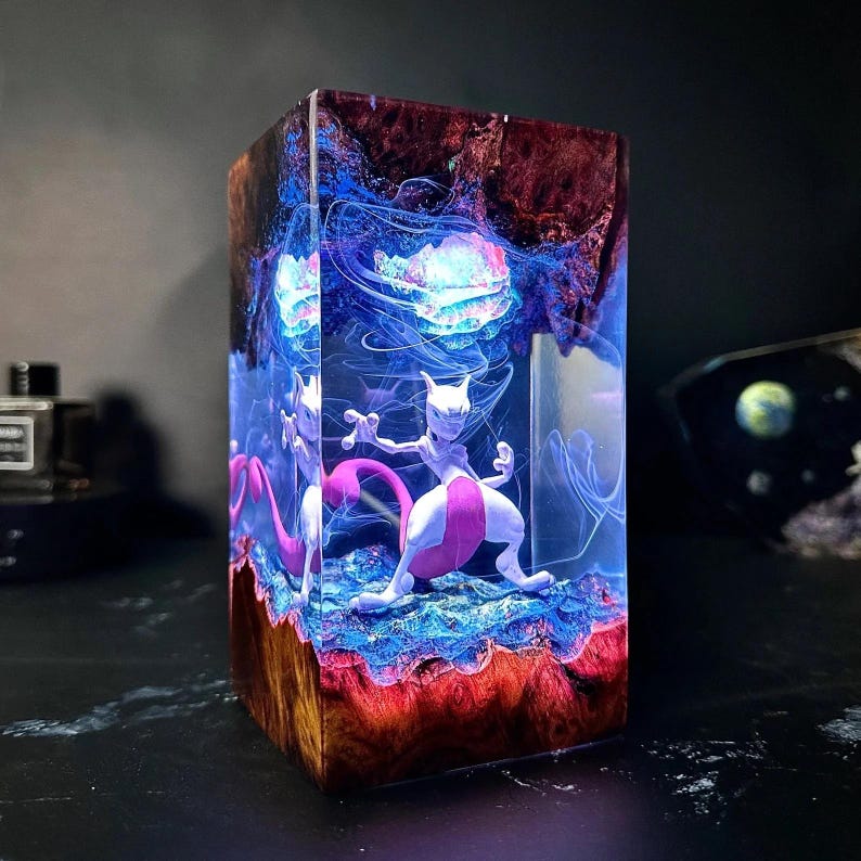 May include: A rectangular resin sculpture featuring a Mewtwo figure. The sculpture has a layered design with red, blue, and purple hues. The Mewtwo figure is white and pink, set against a glowing, cosmic background. The base has a wood-like texture.
