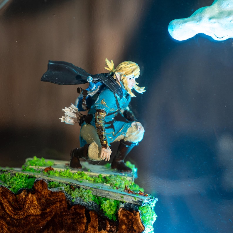 Puede incluir: Figura detallada de un personaje de la serie de videojuegos The Legend of Zelda. La figura est&aacute; en cuclillas, con una t&uacute;nica azul, botas negras y una capa negra. La figura est&aacute; sobre un peque&ntilde;o diorama detallado con musgo verde.