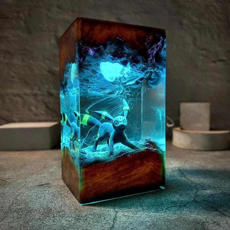 Peut inclure: Lampe rectangulaire en r&eacute;sine avec une figurine Umbreon sombre dans un environnement bleu et violet lumineux. La lampe est encadr&eacute;e de bois brun fonc&eacute;. L'ensemble cr&eacute;e une ambiance mystique et enchanteresse.
