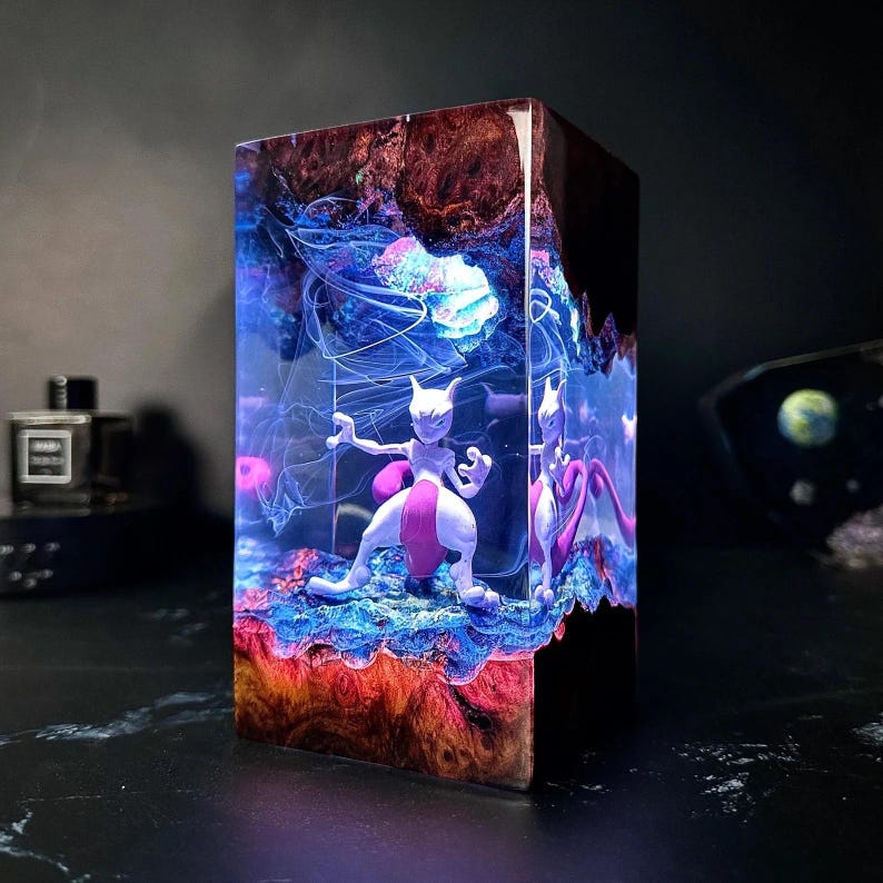 Peut inclure: Sculpture rectangulaire en r&eacute;sine avec une figurine Mewtwo blanche et rose. La sculpture pr&eacute;sente un design en couches avec des teintes bleues, rouges et marron, cr&eacute;ant un effet vibrant et lumineux. Une pi&egrave;ce d&eacute;corative unique.