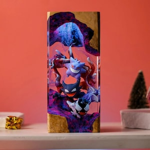 Peut inclure: Une pi&egrave;ce d'art en r&eacute;sine rectangulaire repr&eacute;sentant trois personnages Pok&eacute;mon : Gengar, Haunter et Gastly. Les personnages sont plac&eacute;s sur un fond de r&eacute;sine tourbillonnante violette et bleue, encadr&eacute;s dans un cadre en bois. La pi&egrave;ce est expos&eacute;e sur une surface blanche.