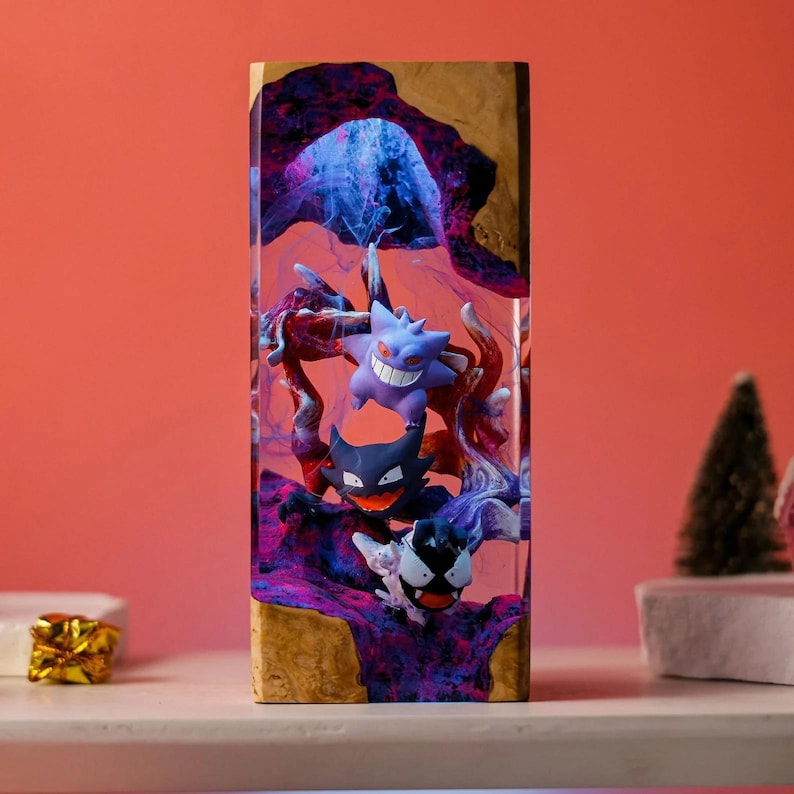 Peut inclure: Une pi&egrave;ce d'art en r&eacute;sine rectangulaire repr&eacute;sentant trois personnages Pok&eacute;mon : Gengar, Haunter et Gastly. Les personnages sont plac&eacute;s sur un fond de r&eacute;sine tourbillonnante violette et bleue, encadr&eacute;s dans un cadre en bois. La pi&egrave;ce est expos&eacute;e sur une surface blanche.