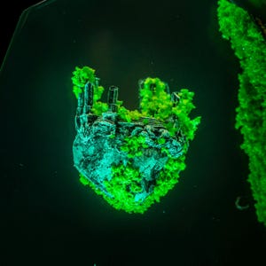 Puede incluir: Una isla de fantas&iacute;a en miniatura con ruinas y exuberante follaje verde. La isla es una mezcla de gris y verde azulado, con vegetaci&oacute;n verde brillante. La escena est&aacute; ambientada sobre un fondo oscuro, creando un efecto dram&aacute;tico.