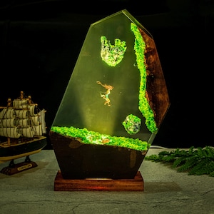 Puede incluir: L&aacute;mpara decorativa de resina con forma geom&eacute;trica. Presenta un paisaje en miniatura con una figura, musgo verde y elementos de madera. La base es de color marr&oacute;n oscuro y descansa sobre un soporte de madera. La paleta de colores es principalmente verde y marr&oacute;n.