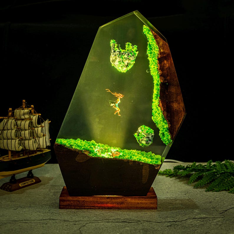 Puede incluir: L&aacute;mpara decorativa de resina con forma geom&eacute;trica. Presenta un paisaje en miniatura con una figura, musgo verde y elementos de madera. La base es de color marr&oacute;n oscuro y descansa sobre un soporte de madera. La paleta de colores es principalmente verde y marr&oacute;n.