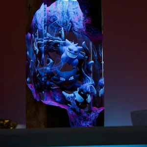 Peut inclure: Une &oelig;uvre d'art en r&eacute;sine repr&eacute;sentant les personnages Pok&eacute;mon bleus Gengar, Haunter et Gastly, sur fond de r&eacute;sine tourbillonnante bleue et violette. L'&oelig;uvre est enferm&eacute;e dans un bloc rectangulaire transparent, &eacute;clair&eacute; par le haut.