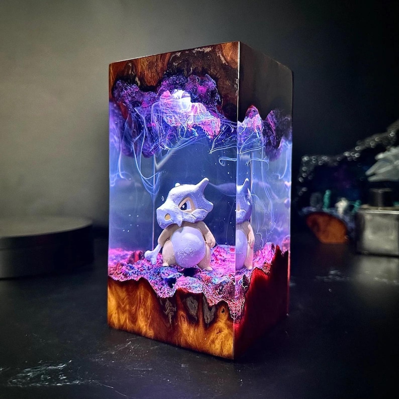 Peut inclure: Une &oelig;uvre d'art en r&eacute;sine rectangulaire repr&eacute;sentant un personnage de Cubone. L'&oelig;uvre a une base en bois avec un design color&eacute; et superpos&eacute;. L'int&eacute;rieur pr&eacute;sente un effet n&eacute;buleux violet et bleu, avec le personnage de Cubone au centre.