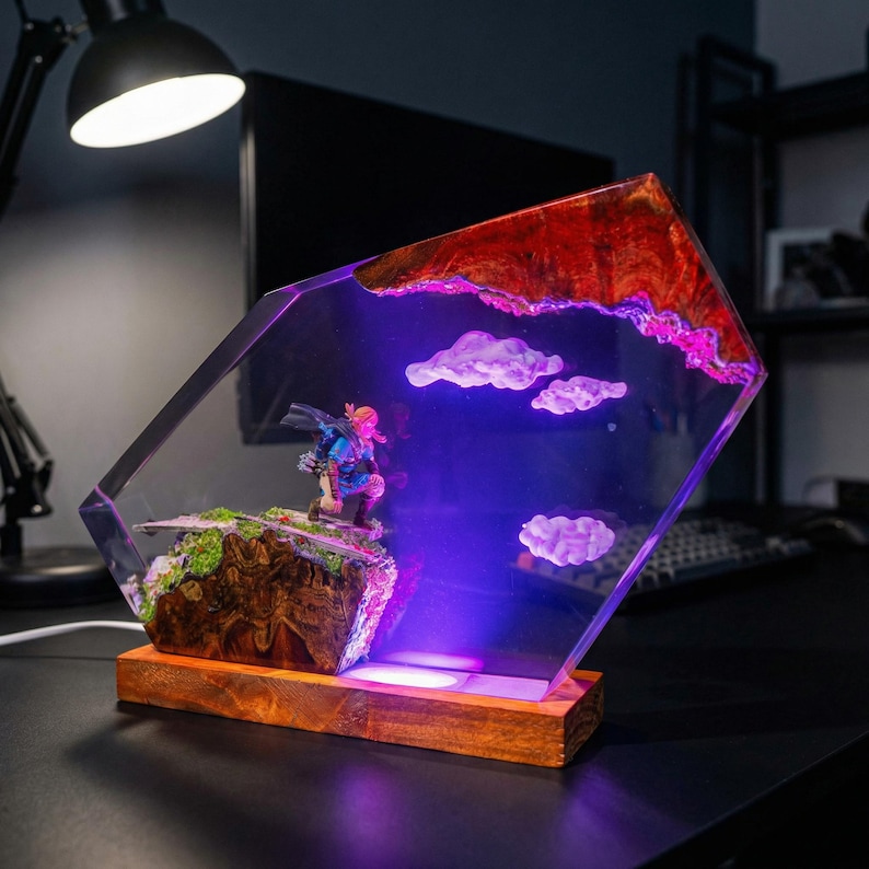 Puede incluir: L&aacute;mpara decorativa con una figura en miniatura sobre un afloramiento rocoso, dentro de un bloque de resina transparente y angular. La secci&oacute;n superior tiene un dise&ntilde;o rojo y marr&oacute;n, con formas de nubes blancas. La base es de madera.