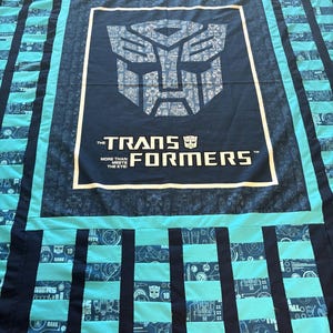 Puede incluir: Un edredón hecho a mano con un gran cuadrado azul marino central que muestra el logotipo y el texto de Transformers. El edredón tiene un patrón de rayas turquesa y azul marino. El texto dice "THE TRANSFORMERS MORE THAN MEETS THE EYE!"