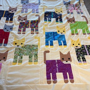 May include: A handmade quilt featuring a repeating pattern of stylized cats. Each cat is a different color and pattern, including floral, polka dot, and solid colors. The quilt is on a cream-colored background.