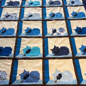 Peut inclure: Une courtepointe faite à la main avec un motif répété de silhouettes de chats endormis dans diverses nuances de bleu. Les chats sont placés sur un fond crème, encadrés de bordures marron et bleu. Le bord de la courtepointe a une bordure bleu foncé à pois.