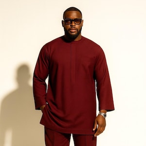 May include: A burgundy two-piece suit, comprising a long-sleeved tunic with a single chest pocket and matching trousers. The outfit is complemented by brown leather shoes and a silver wristwatch. The background is plain white.