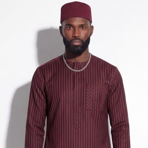 May include: A burgundy two-piece suit with vertical white pinstripes. The long-sleeved shirt has a single pocket and a mandarin collar. The matching pants are straight-legged. A burgundy cap and a silver chain complete the outfit.