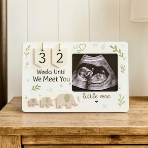 Puede incluir: Marco de anuncio de embarazo de madera blanca con una imagen de ultrasonido. El marco tiene el texto "32 semanas hasta que te conozcamos" y "pequeño". Los recortes decorativos de elefantes y los acentos de hojas completan el diseño.