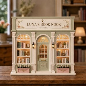 Könnte beinhalten: Ein detailliertes Miniaturmodell von Luna's Book Nook, einer Buchhandlung mit grüner und weißer Fassade. Das Schild lautet "Luna's Book Nook" mit floralen Akzenten. Das Modell verfügt über gewölbte Fenster, eine zentrale Tür und Regale mit winzigen Büchern.