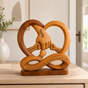 Könnte beinhalten: Holzskulptur in Herzform mit ineinander verschlungenen Händen. Das Wort "Daddy" ist auf der größten Hand eingraviert, mit "Emma", "Noah" und "Liam" auf kleineren Händen. Auf dem Sockel steht: "Für die Welt bist du ein Papa, aber für unsere Familie bist du die Welt."