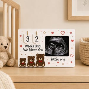May include: A white rectangular wooden frame featuring a black and white ultrasound image, two hanging number tags displaying 3 and 2, and the text "Weeks Until We Meet You" and "little one". The frame is decorated with three brown bear icons.