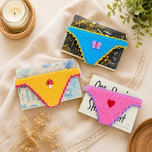 May include: Three crochet underwear-shaped bookmarks in blue, yellow, and pink. The blue bookmark has a butterfly, the yellow has a mushroom, and the pink has a heart. Each bookmark is placed on top of a book.