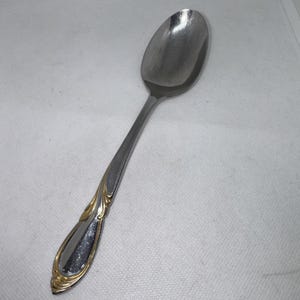 May include: A stainless steel spoon with a polished finish. The handle features a decorative gold-colored design. The spoon is designed for serving and eating. The spoon is on a white surface.